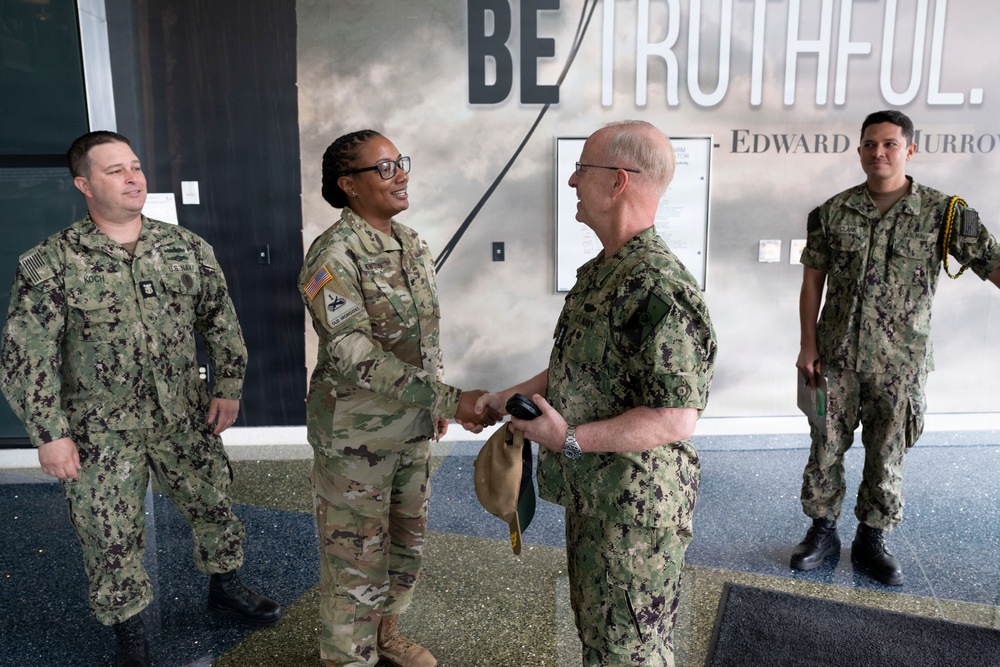Four military personnel are gathered in front of a building entrance featuring a 'BE TRUTHFUL' quote attributed to Edward R. Murrow. A U.S. Army soldier in OCP (Operational Camouflage Pattern) uniform is shaking hands with a senior U.S. Navy officer in NWU Type III uniform, suggesting a formal greeting or meeting. Two additional Navy personnel in NWU Type III uniforms are present, one on each side.