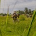 A soldier in camouflage uniform is kneeling in tall grass, aiming a rifle in a tropical environment. Several other military personnel are visible in the background across an open field. The setting appears to be a training exercise or tactical operation in a tropical region, evidenced by palm trees and lush vegetation.