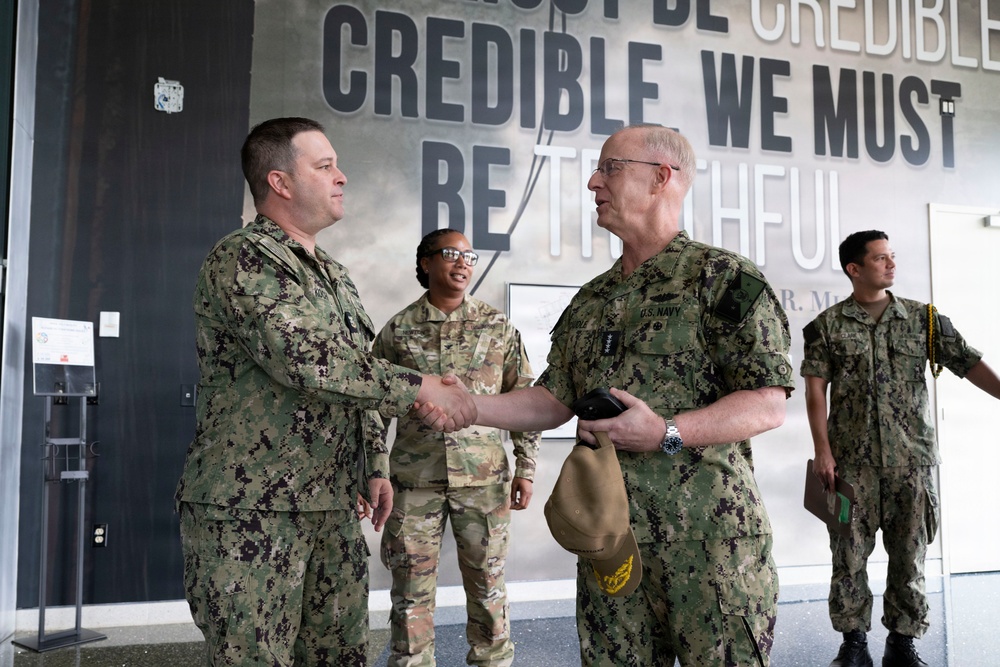 Four military personnel are gathered in what appears to be a modern facility or headquarters building. Two individuals in U.S. Navy NWU Type III (digital camouflage) uniforms are shaking hands in the foreground, while two others in different camouflage patterns stand in the background. A large motivational text mural reading 'MUST BE CREDIBLE, CREDIBLE WE MUST BE TRUTHFUL' is prominently displayed on the wall behind them.