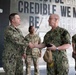 Four military personnel are gathered in what appears to be a modern facility or headquarters building. Two individuals in U.S. Navy NWU Type III (digital camouflage) uniforms are shaking hands in the foreground, while two others in different camouflage patterns stand in the background. A large motivational text mural reading 'MUST BE CREDIBLE, CREDIBLE WE MUST BE TRUTHFUL' is prominently displayed on the wall behind them.