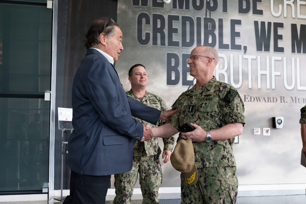 A civilian in a blue suit is shaking hands with a senior U.S. Navy officer in digital camouflage (NWU Type III) uniform, while a second Navy enlisted sailor stands in the background observing. The setting appears to be an official military facility, with a large wall mural quoting Edward R. Murrow: 'We must be credible, we must be truthful.' The interaction suggests a formal greeting or official visit.
