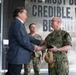 A civilian in a blue suit is shaking hands with a senior U.S. Navy officer in digital camouflage (NWU Type III) uniform, while a second Navy enlisted sailor stands in the background observing. The setting appears to be an official military facility, with a large wall mural quoting Edward R. Murrow: 'We must be credible, we must be truthful.' The interaction suggests a formal greeting or official visit.