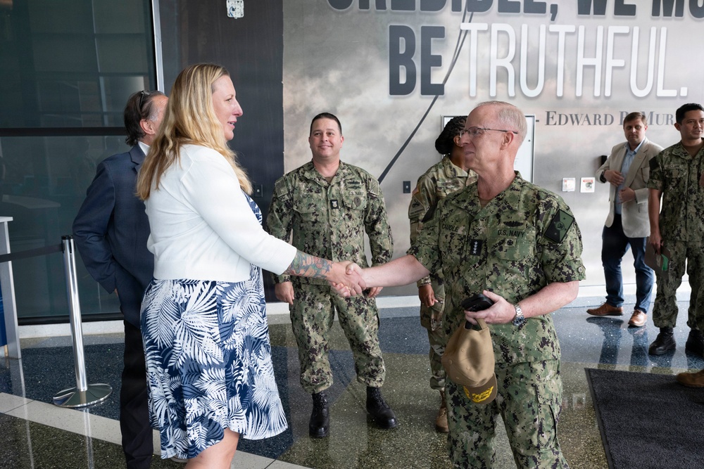 A civilian woman in a white blazer and floral skirt is shaking hands with a senior U.S. Navy officer in digital camouflage uniform. The meeting appears to be a formal greeting or reception, taking place in what appears to be a modern building lobby. Several other military personnel in U.S. Navy uniforms are present in the background observing the exchange.