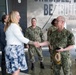 A civilian woman in a white blazer and floral skirt is shaking hands with a senior U.S. Navy officer in digital camouflage uniform. The meeting appears to be a formal greeting or reception, taking place in what appears to be a modern building lobby. Several other military personnel in U.S. Navy uniforms are present in the background observing the exchange.