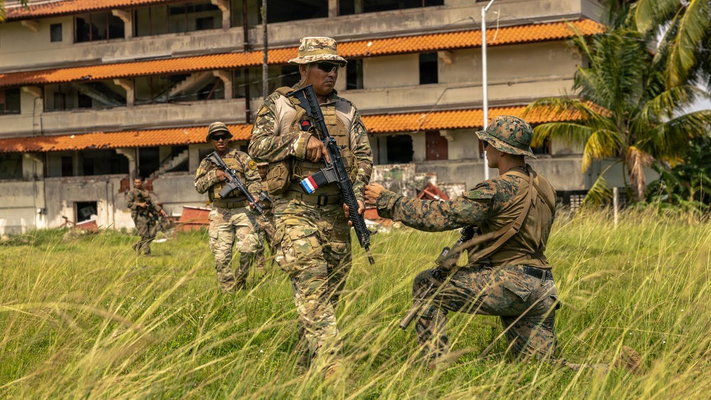 Military personnel in camouflage uniforms conduct what appears to be a joint training exercise in an outdoor area near a deteriorating multi-story building. At least four soldiers are visible, with one kneeling and appearing to interact with a standing soldier who bears a flag patch on his uniform. The tropical vegetation and architecture suggest a location in a warm, humid region.