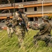 Military personnel in camouflage uniforms conduct what appears to be a joint training exercise in an outdoor area near a deteriorating multi-story building. At least four soldiers are visible, with one kneeling and appearing to interact with a standing soldier who bears a flag patch on his uniform. The tropical vegetation and architecture suggest a location in a warm, humid region.