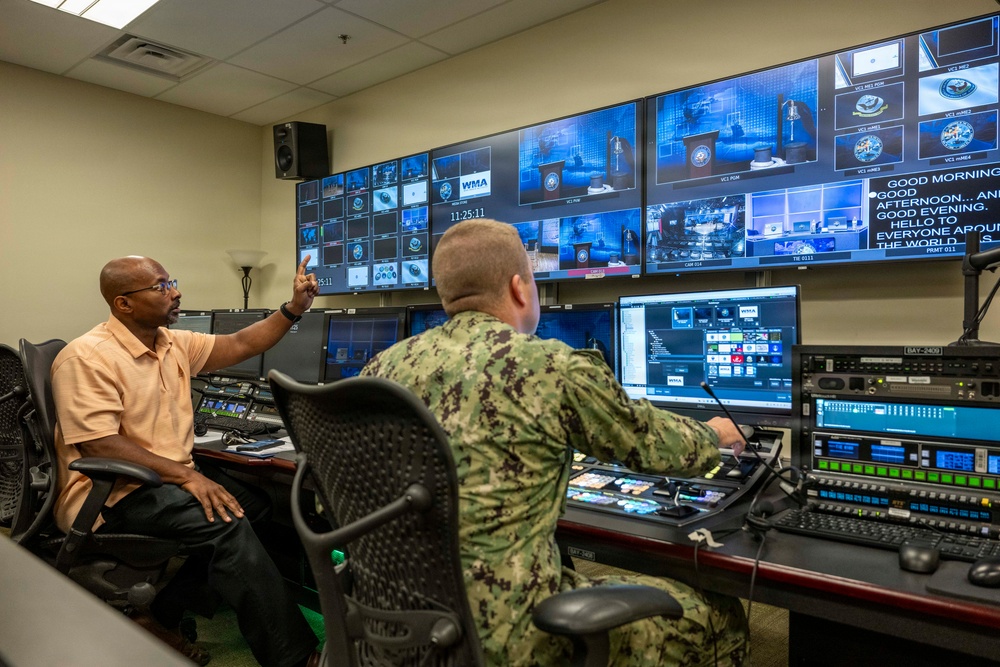 Two individuals are working in a professional broadcast control room equipped with multiple monitors and production equipment. One person in civilian attire is gesturing toward the video wall while a military personnel member in camouflage uniform operates the production console. The room displays numerous video feeds, a timestamp reading 11:25:11, and text reading 'WMA' on several screens.