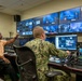 Two individuals are working in a professional broadcast control room equipped with multiple monitors and production equipment. One person in civilian attire is gesturing toward the video wall while a military personnel member in camouflage uniform operates the production console. The room displays numerous video feeds, a timestamp reading 11:25:11, and text reading 'WMA' on several screens.