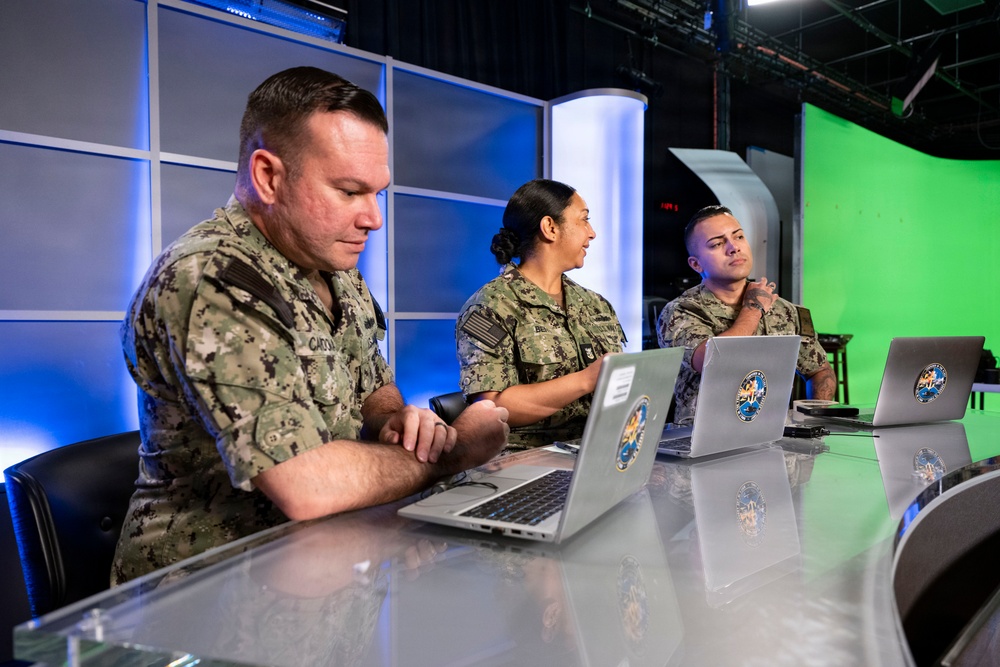Three U.S. Navy personnel in NWU Type III (digital camouflage) uniforms are seated at a reflective table in what appears to be a broadcast or media studio. Each person has a laptop computer with a circular military unit insignia sticker visible on the lid. The setting includes blue accent lighting, a green screen backdrop, and studio infrastructure visible in the background.