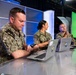 Three U.S. Navy personnel in NWU Type III (digital camouflage) uniforms are seated at a reflective table in what appears to be a broadcast or media studio. Each person has a laptop computer with a circular military unit insignia sticker visible on the lid. The setting includes blue accent lighting, a green screen backdrop, and studio infrastructure visible in the background.