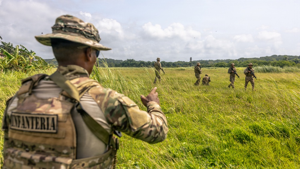 A military training exercise is taking place in an open grassy field with tropical vegetation in the background. A soldier in the foreground wearing a patch labeled 'INFANTERIA' and a boonie hat is gesturing toward a group of approximately four soldiers moving through the field. The setting appears to be a tropical or subtropical environment, suggesting a joint or multinational training operation.