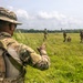 A military training exercise is taking place in an open grassy field with tropical vegetation in the background. A soldier in the foreground wearing a patch labeled 'INFANTERIA' and a boonie hat is gesturing toward a group of approximately four soldiers moving through the field. The setting appears to be a tropical or subtropical environment, suggesting a joint or multinational training operation.