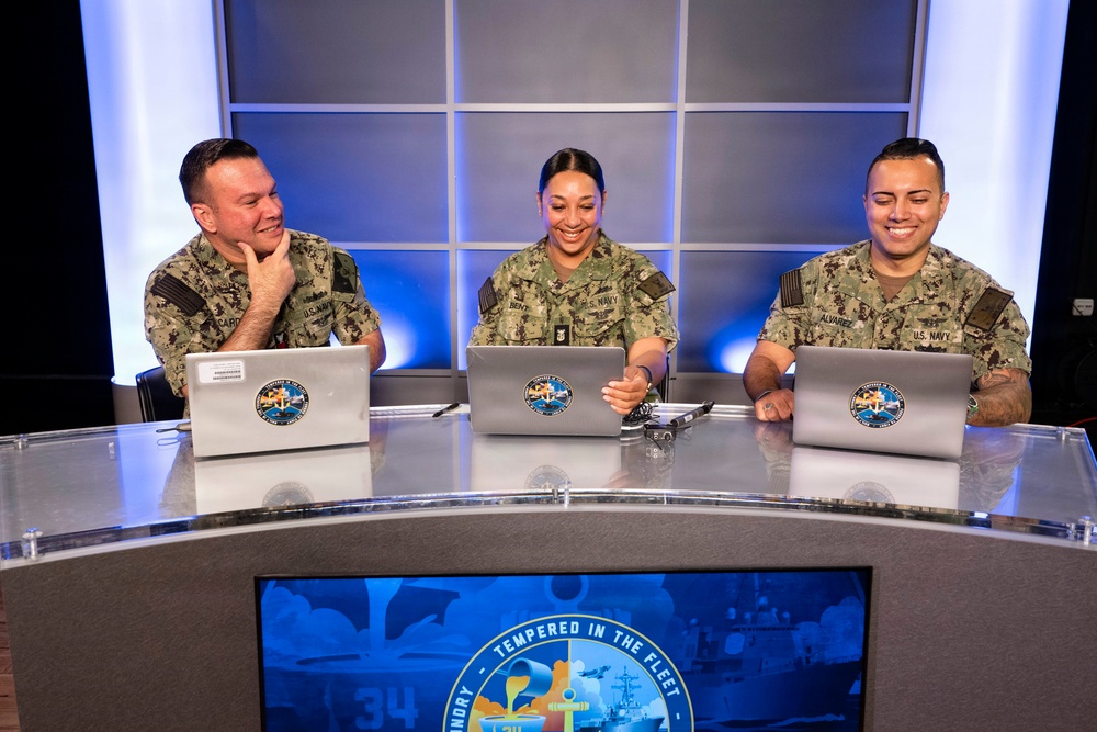 Three U.S. Navy personnel in NWU (Navy Working Uniform) camouflage are seated at a curved broadcast desk with laptops bearing official Navy insignia stickers. They appear to be participating in a televised or recorded Navy production, as evidenced by the professional studio lighting and a monitor displaying 'Tempered in the Fleet' branding. All three individuals are smiling and appear engaged in a positive, collaborative setting.