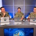 Three U.S. Navy personnel in NWU (Navy Working Uniform) camouflage are seated at a curved broadcast desk with laptops bearing official Navy insignia stickers. They appear to be participating in a televised or recorded Navy production, as evidenced by the professional studio lighting and a monitor displaying 'Tempered in the Fleet' branding. All three individuals are smiling and appear engaged in a positive, collaborative setting.