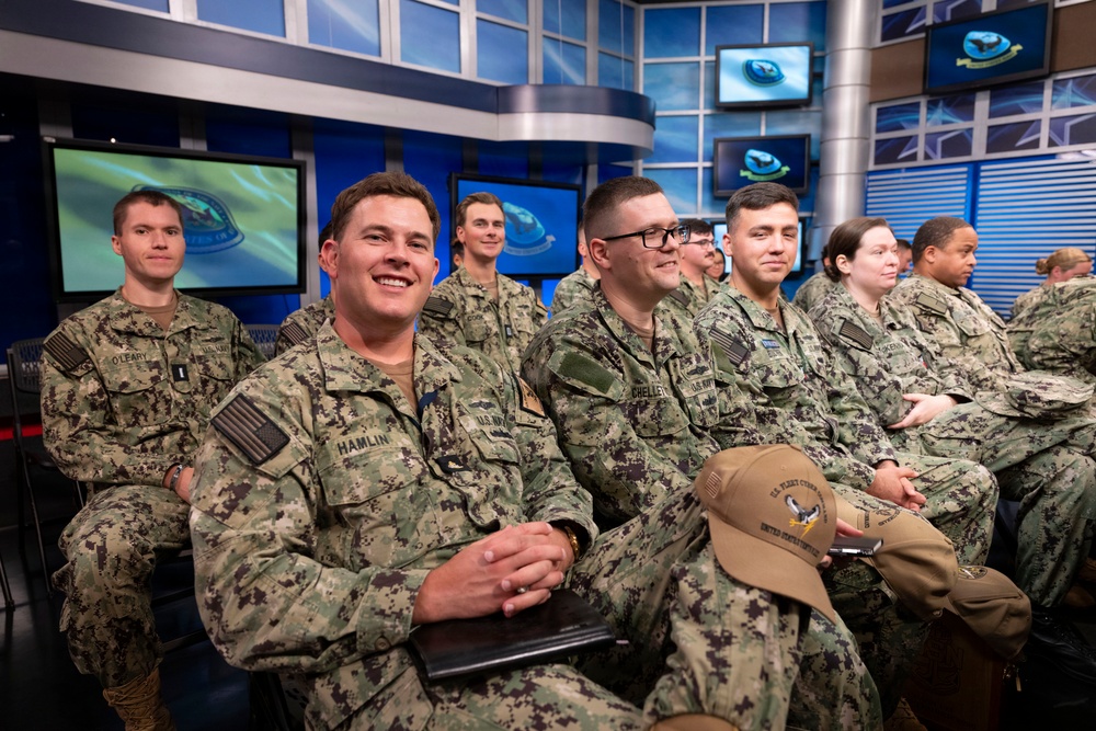 A group of U.S. Navy personnel in NWU Type III (digital camouflage) uniforms are seated in what appears to be a military broadcast studio or media facility. Multiple monitors displaying what appears to be a Navy logo/emblem are visible in the background. The personnel appear relaxed and engaged, with several smiling.