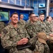 A group of U.S. Navy personnel in NWU Type III (digital camouflage) uniforms are seated in what appears to be a military broadcast studio or media facility. Multiple monitors displaying what appears to be a Navy logo/emblem are visible in the background. The personnel appear relaxed and engaged, with several smiling.