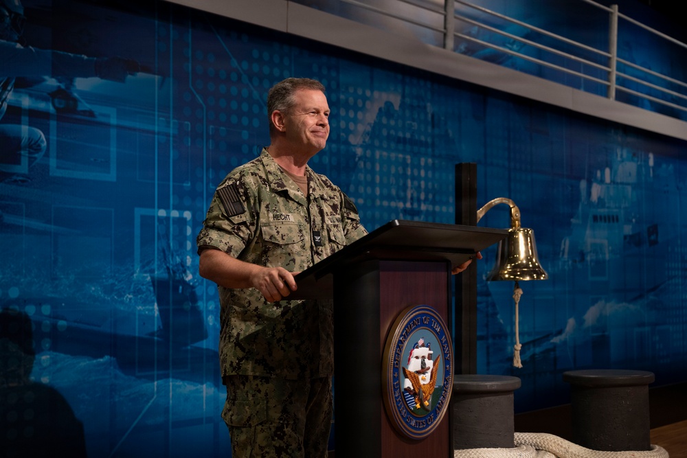 A U.S. Navy officer wearing digital camouflage NWU (Navy Working Uniform) stands at a podium bearing the Department of the Navy seal. The individual, whose name tape reads 'HECHT,' appears to be delivering a speech or address at an official Navy event. A traditional brass ship's bell is mounted to the right of the podium, and a blue digital world map backdrop is visible behind the speaker.
