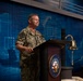 A U.S. Navy officer wearing digital camouflage NWU (Navy Working Uniform) stands at a podium bearing the Department of the Navy seal. The individual, whose name tape reads 'HECHT,' appears to be delivering a speech or address at an official Navy event. A traditional brass ship's bell is mounted to the right of the podium, and a blue digital world map backdrop is visible behind the speaker.