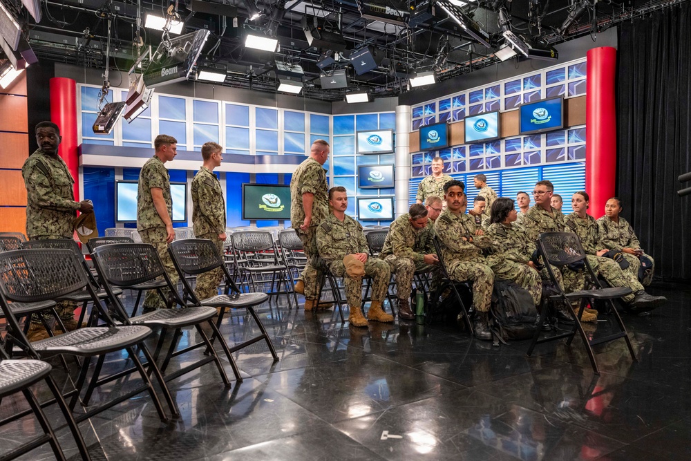 A group of military personnel in U.S. Navy digital camouflage uniforms (NWU Type I) are gathered inside what appears to be a professional television broadcast studio. The studio features multiple monitors displaying a logo, professional lighting rigs, and a polished studio set with red pillars and blue backdrop panels. Personnel are both seated in folding chairs and standing, suggesting a briefing, tour, or event is underway.