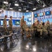 A group of military personnel in U.S. Navy digital camouflage uniforms (NWU Type I) are gathered inside what appears to be a professional television broadcast studio. The studio features multiple monitors displaying a logo, professional lighting rigs, and a polished studio set with red pillars and blue backdrop panels. Personnel are both seated in folding chairs and standing, suggesting a briefing, tour, or event is underway.