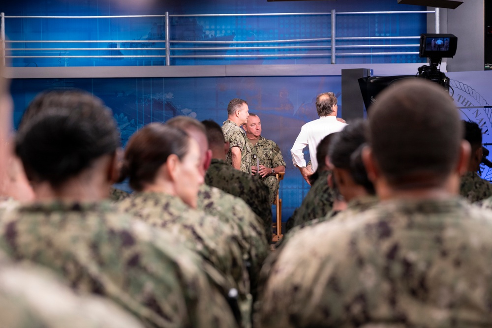 A military briefing or town hall event is taking place in what appears to be a media studio or briefing room with a blue backdrop. Multiple personnel in U.S. Navy NWU (Naval Working Uniform) camouflage are seated as an audience, facing two uniformed individuals and one civilian in a white shirt at the front. The setting includes professional camera equipment and a stylized blue background with map graphics.