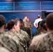 A military briefing or town hall event is taking place in what appears to be a media studio or briefing room with a blue backdrop. Multiple personnel in U.S. Navy NWU (Naval Working Uniform) camouflage are seated as an audience, facing two uniformed individuals and one civilian in a white shirt at the front. The setting includes professional camera equipment and a stylized blue background with map graphics.