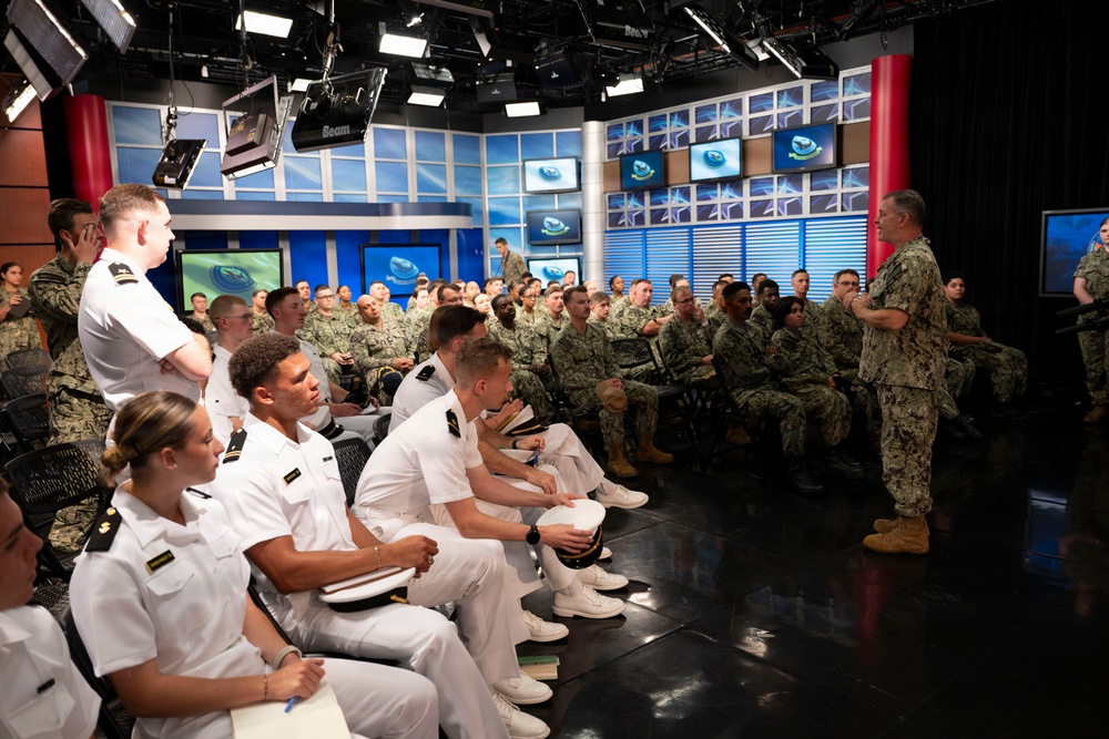 A senior military officer in Navy Working Uniform (NWU) addresses a large audience of naval personnel in what appears to be a broadcast studio or media center. The audience includes both officers in white dress uniforms and enlisted personnel in camouflage NWU Type III uniforms. The setting features professional broadcast lighting, multiple monitors, and studio equipment consistent with a military media or communications facility.