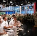 A senior military officer in Navy Working Uniform (NWU) addresses a large audience of naval personnel in what appears to be a broadcast studio or media center. The audience includes both officers in white dress uniforms and enlisted personnel in camouflage NWU Type III uniforms. The setting features professional broadcast lighting, multiple monitors, and studio equipment consistent with a military media or communications facility.