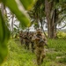 A column of armed military personnel in camouflage uniforms patrol single-file along a narrow jungle trail surrounded by dense tropical vegetation including palm trees and broad-leafed plants. The lead soldier wears a boonie hat and MultiCam/OCP-pattern uniform while carrying what appears to be an M4-style carbine rifle. The group behind him appears to wear a mix of camouflage patterns, suggesting a possible joint or multinational training exercise.