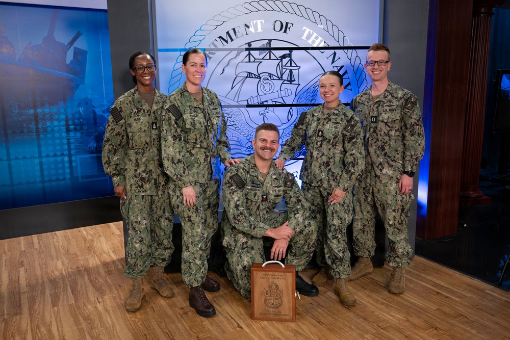 Five U.S. Navy personnel in NWU Type III (woodland camouflage) uniforms pose for a group photo in what appears to be a media studio or briefing room. Four individuals stand while one kneels in the center holding a wooden award plaque bearing the U.S. Navy anchor insignia. The backdrop displays the Department of the Navy seal, suggesting an official recognition or award ceremony.