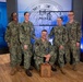 Five U.S. Navy personnel in NWU Type III (woodland camouflage) uniforms pose for a group photo in what appears to be a media studio or briefing room. Four individuals stand while one kneels in the center holding a wooden award plaque bearing the U.S. Navy anchor insignia. The backdrop displays the Department of the Navy seal, suggesting an official recognition or award ceremony.