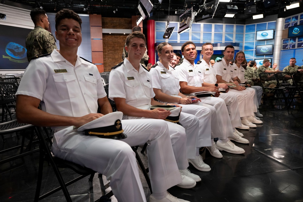 A group of naval cadets or midshipmen in white dress uniforms are seated in a row at what appears to be a broadcast studio or media facility. They are smiling and appear to be attending a formal event or presentation. The background shows a professional studio environment with screens, lighting rigs, and additional military personnel in camouflage uniforms.