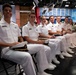 A group of naval cadets or midshipmen in white dress uniforms are seated in a row at what appears to be a broadcast studio or media facility. They are smiling and appear to be attending a formal event or presentation. The background shows a professional studio environment with screens, lighting rigs, and additional military personnel in camouflage uniforms.