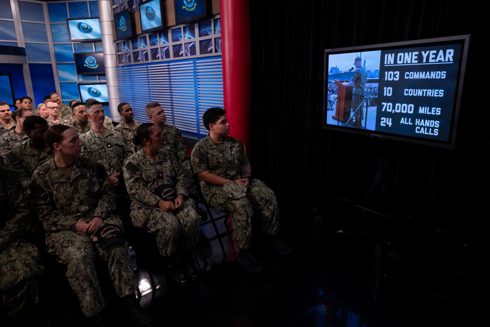 A group of U.S. Navy personnel in digital camouflage uniforms are seated in what appears to be a broadcast studio or media center, watching a large screen displaying statistics. The screen shows an infographic titled 'IN ONE YEAR' listing accomplishments: 103 Commands, 10 Countries, 70,000 Miles, and 24 All Hands Calls. The setting features multiple monitors displaying a naval insignia logo on the walls behind the audience.