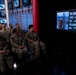 A group of U.S. Navy personnel in digital camouflage uniforms are seated in what appears to be a broadcast studio or media center, watching a large screen displaying statistics. The screen shows an infographic titled 'IN ONE YEAR' listing accomplishments: 103 Commands, 10 Countries, 70,000 Miles, and 24 All Hands Calls. The setting features multiple monitors displaying a naval insignia logo on the walls behind the audience.