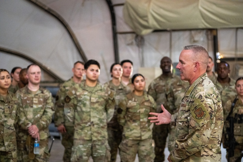 A senior military officer in OCP (Operational Camouflage Pattern) uniform is addressing a group of approximately 20 military personnel inside what appears to be a large military tent or temporary structure. The speaker, positioned on the right side of the frame, is gesturing with his hands while speaking to the assembled troops who are standing at ease. The setting suggests a briefing, commander's call, or unit address at a forward operating location.