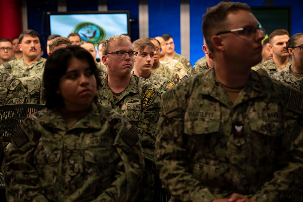 A large group of U.S. Navy personnel in Navy Working Uniform (NWU) camouflage are gathered in what appears to be a briefing room or auditorium. The service members are seated and standing in rows, appearing to watch or listen to a presentation. A screen in the background displays what appears to be a U.S. Department of Defense or military command seal.