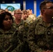 A large group of U.S. Navy personnel in Navy Working Uniform (NWU) camouflage are gathered in what appears to be a briefing room or auditorium. The service members are seated and standing in rows, appearing to watch or listen to a presentation. A screen in the background displays what appears to be a U.S. Department of Defense or military command seal.