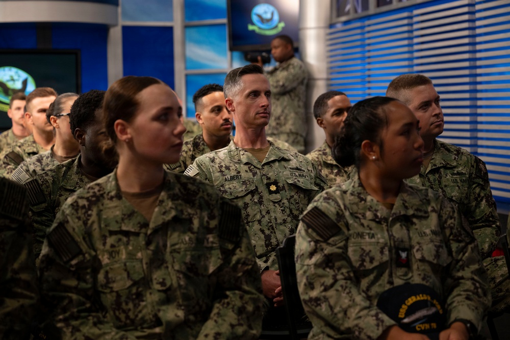 A group of U.S. Navy personnel in NWU Type III (Navy Working Uniform) camouflage are gathered in what appears to be a briefing room or media studio with blue and teal decor. The personnel appear to be attentively watching or listening to a presentation. One individual in the center is identifiable by a name tape reading 'ALBERT' and wears a gold oak leaf insignia indicating the rank of Lieutenant Commander or Major.