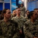 A group of U.S. Navy personnel in NWU Type III (Navy Working Uniform) camouflage are gathered in what appears to be a briefing room or media studio with blue and teal decor. The personnel appear to be attentively watching or listening to a presentation. One individual in the center is identifiable by a name tape reading 'ALBERT' and wears a gold oak leaf insignia indicating the rank of Lieutenant Commander or Major.