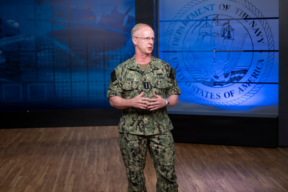 A U.S. Navy officer in digital camouflage (NWU Type III) uniform stands before a Department of the Navy seal backdrop in what appears to be a formal briefing or presentation setting. The officer's name tape reads 'CAUDLE' and rank insignia is visible on the uniform. The setting features professional studio-quality lighting with a blue-illuminated background.