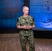 A U.S. Navy officer in digital camouflage (NWU Type III) uniform stands before a Department of the Navy seal backdrop in what appears to be a formal briefing or presentation setting. The officer's name tape reads 'CAUDLE' and rank insignia is visible on the uniform. The setting features professional studio-quality lighting with a blue-illuminated background.