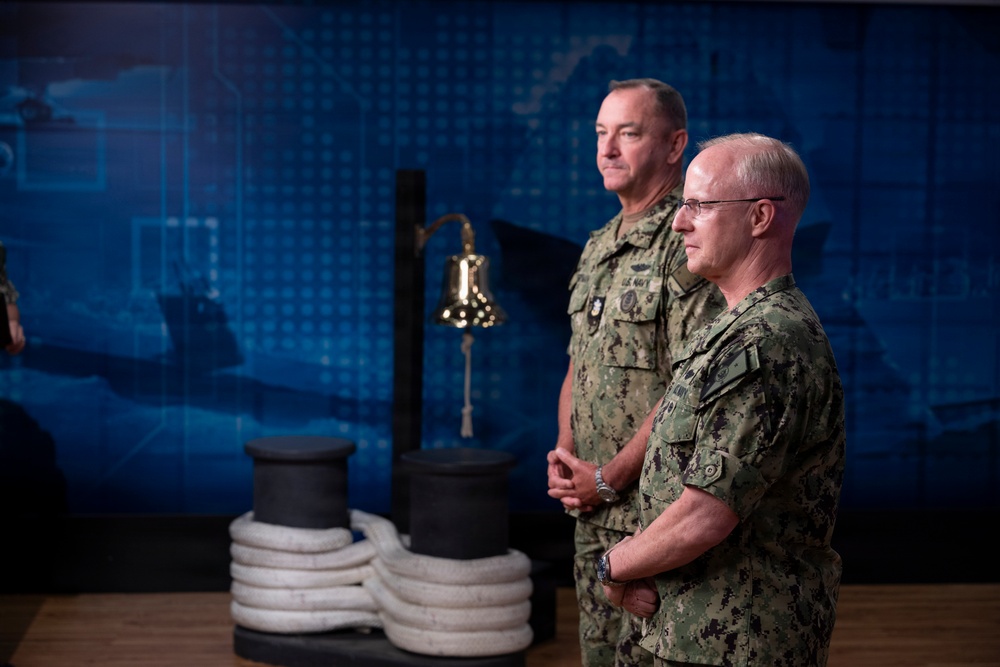 Two U.S. Navy personnel in NWU (Navy Working Uniform) Type III camouflage stand at attention in what appears to be a formal military setting or briefing room. Both individuals have visible U.S. Navy name tapes and insignia on their uniforms. The backdrop features a blue digital world map display along with nautical props including a ship's bell, mooring bollard, and coiled rope.