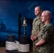 Two U.S. Navy personnel in NWU (Navy Working Uniform) Type III camouflage stand at attention in what appears to be a formal military setting or briefing room. Both individuals have visible U.S. Navy name tapes and insignia on their uniforms. The backdrop features a blue digital world map display along with nautical props including a ship's bell, mooring bollard, and coiled rope.