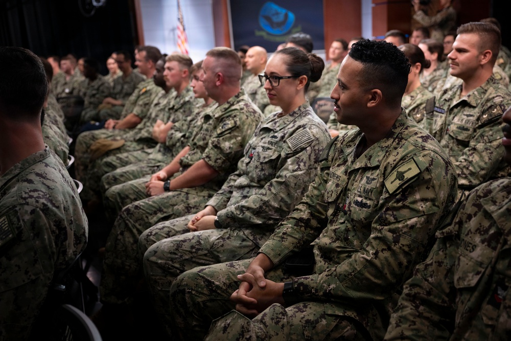 A large group of U.S. Navy personnel in NWU Type III (green camouflage) uniforms are seated in rows, appearing to attend a briefing, ceremony, or official presentation. The audience is mixed in gender and ethnicity, all wearing matching military uniforms. The setting appears to be an indoor auditorium or conference hall with an American flag and an official logo/banner visible in the background.