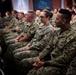 A large group of U.S. Navy personnel in NWU Type III (green camouflage) uniforms are seated in rows, appearing to attend a briefing, ceremony, or official presentation. The audience is mixed in gender and ethnicity, all wearing matching military uniforms. The setting appears to be an indoor auditorium or conference hall with an American flag and an official logo/banner visible in the background.
