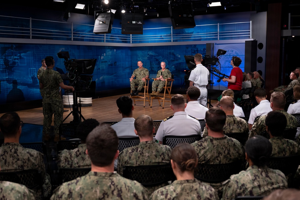 A formal military briefing or town hall event is taking place in a professional broadcast studio setting. Two senior Navy officers in camouflage uniforms are seated on chairs on a raised stage, addressing an audience of Navy personnel. The event appears to be a recorded or live broadcast, with professional camera equipment and crew visible.