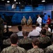 A formal military briefing or town hall event is taking place in a professional broadcast studio setting. Two senior Navy officers in camouflage uniforms are seated on chairs on a raised stage, addressing an audience of Navy personnel. The event appears to be a recorded or live broadcast, with professional camera equipment and crew visible.