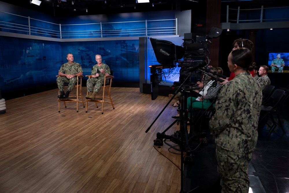 Two U.S. Navy personnel in digital camouflage uniforms are seated on wooden chairs in a professional broadcast studio, being filmed for what appears to be an official military media production. A camera operator in Navy uniform operates a professional broadcast camera with a teleprompter attached, while several civilian and military observers watch from the side. The studio features a blue digital backdrop with naval imagery and professional lighting equipment.