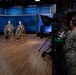 Two U.S. Navy personnel in digital camouflage uniforms are seated on wooden chairs in a professional broadcast studio, being filmed for what appears to be an official military media production. A camera operator in Navy uniform operates a professional broadcast camera with a teleprompter attached, while several civilian and military observers watch from the side. The studio features a blue digital backdrop with naval imagery and professional lighting equipment.
